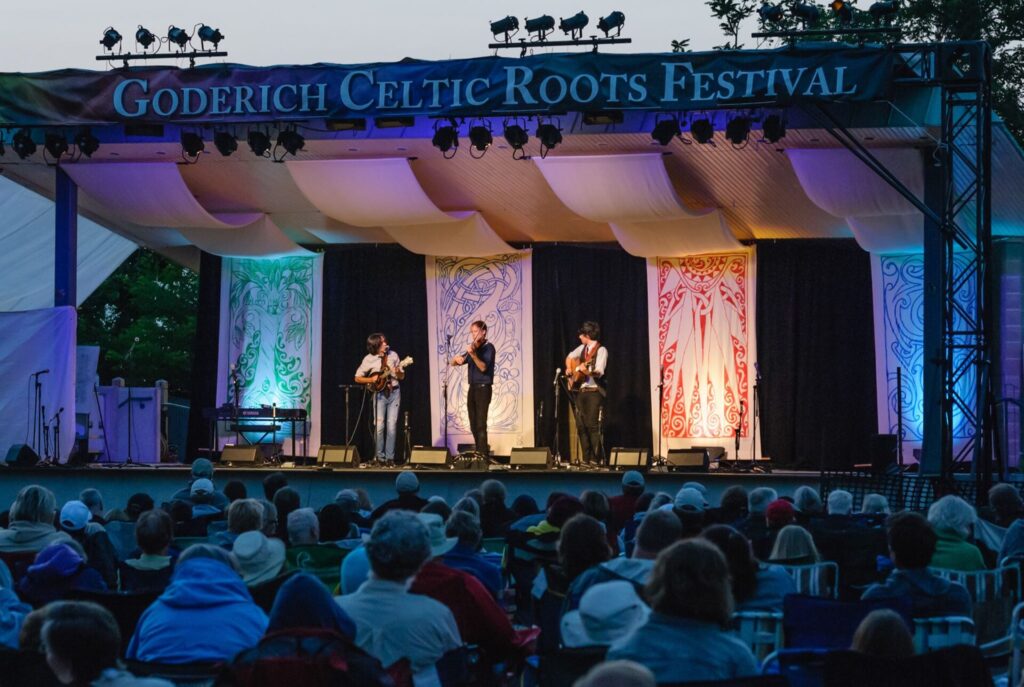 three performers on stage in front of people in a crowd, goderich celtic roots festival sign above stage